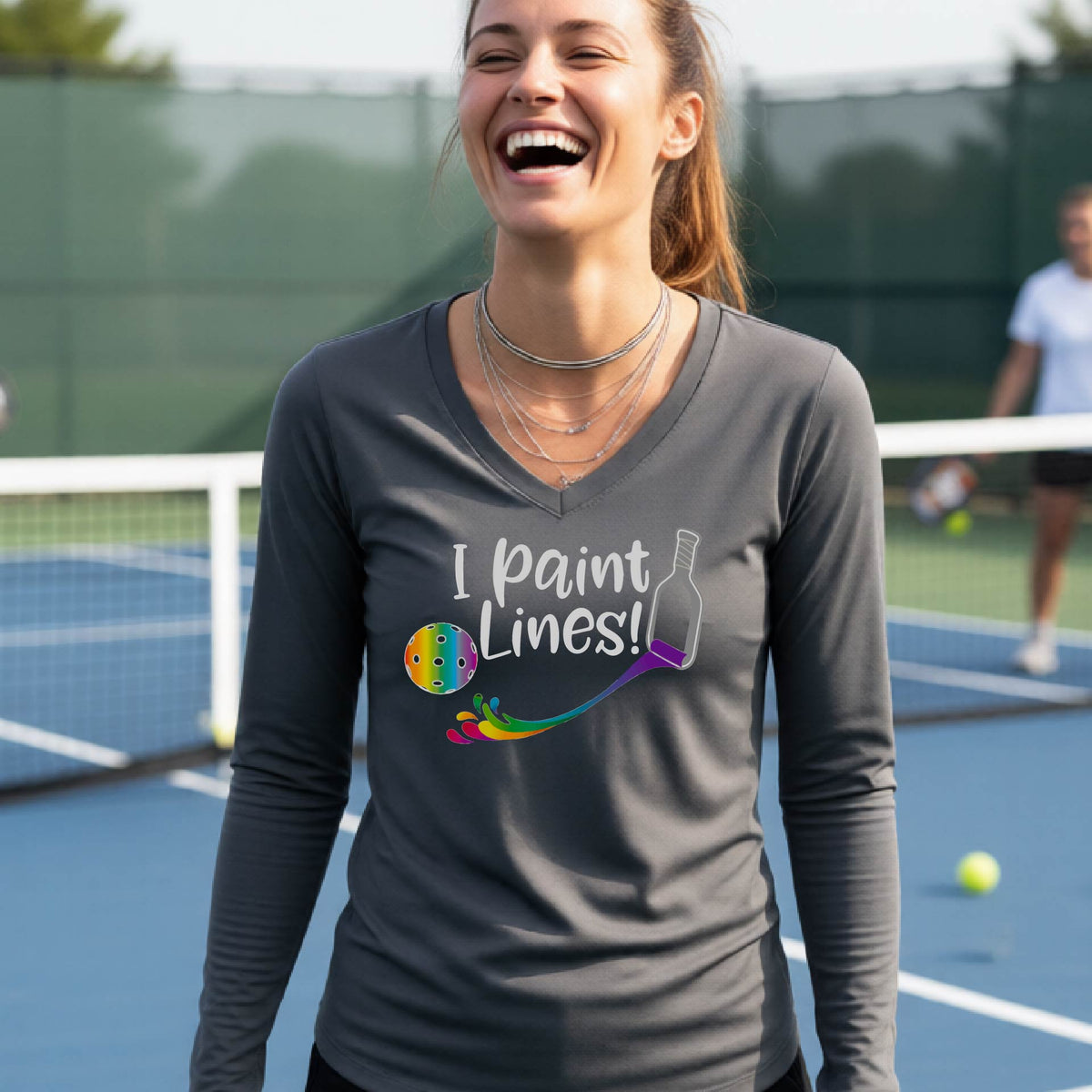 A close-up of a woman wearing a coral dark gray long-sleeve v-neck t-shirt with a graphic on the front that has white text "I PAINT LINES'  with a white pickleball paddle painting multi-colored lines leading a mutli-colored pickleball.