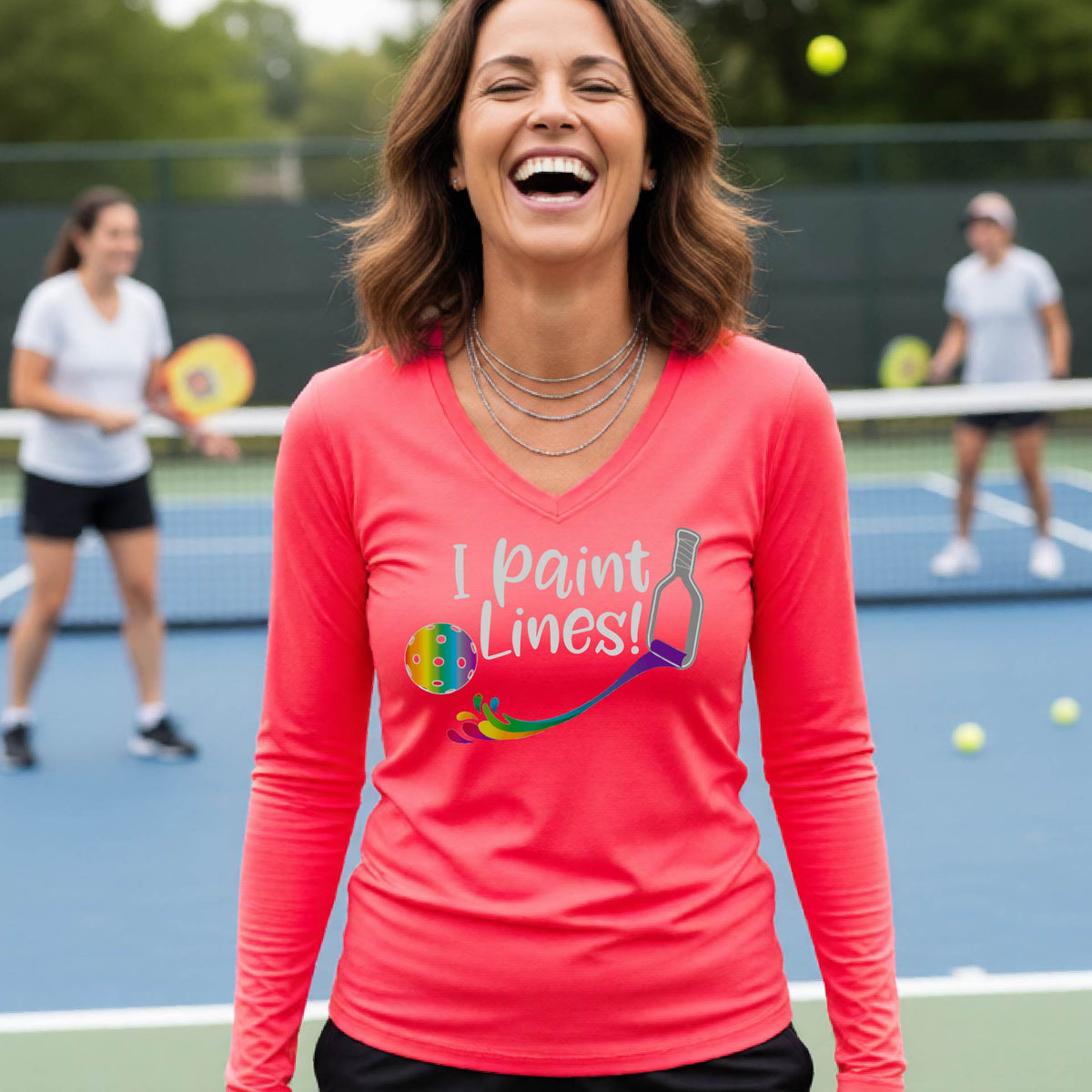 A close-up of a woman wearing a coral pink long-sleeve v-neck t-shirt with a graphic on the front that has white text "I PAINT LINES'  with a white pickleball paddle painting multi-colored lines leading a mutli-colored pickleball.