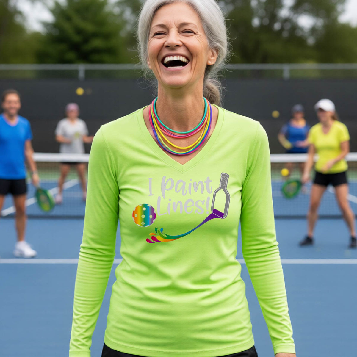 A close-up of a woman wearing a coral lime green long-sleeve v-neck t-shirt with a graphic on the front that has white text "I PAINT LINES'  with a white pickleball paddle painting multi-colored lines leading a mutli-colored pickleball.