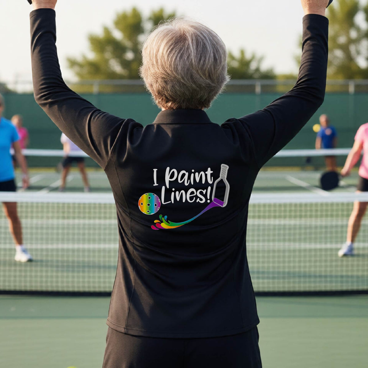 A woman standing on a pickleball court seen from behind, wearing a black long-sleeve shirt with the phrase 'I paint Lines!' and a pickelball paddle graphic printed on the back including a multi-colored pickleball. 