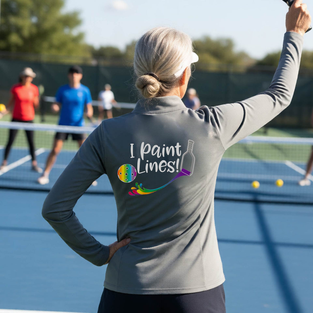 A woman standing on a pickleball court seen from behind, wearing a gray long-sleeve shirt with the phrase 'I paint Lines!' and a pickelball paddle graphic printed on the back including a multi-colored pickleball. 