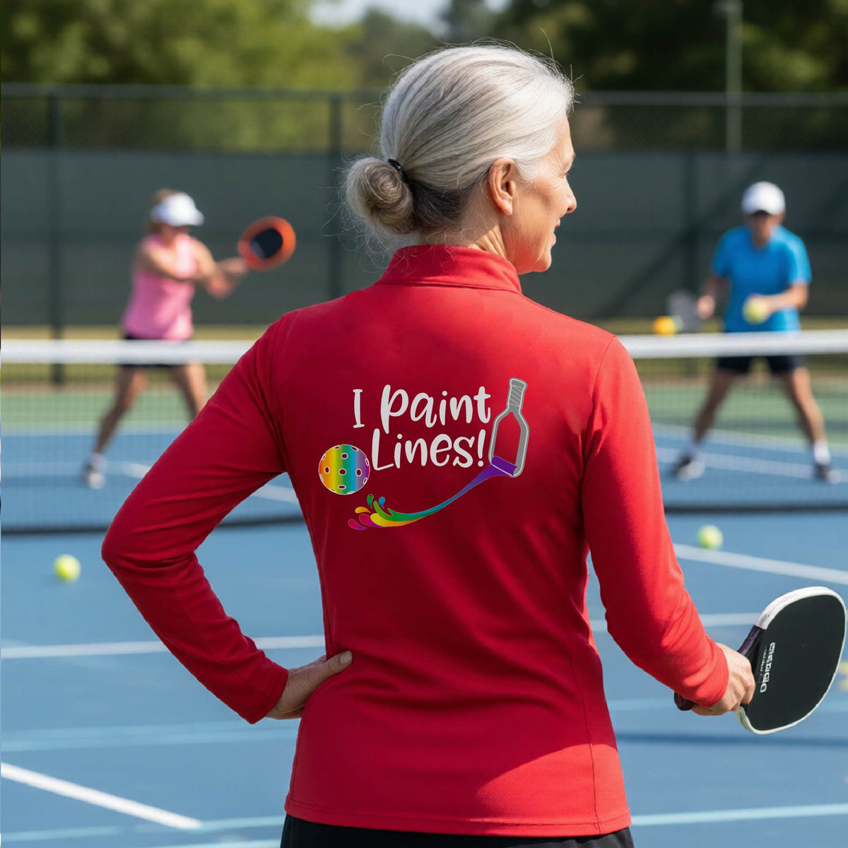 A woman standing on a pickleball court seen from behind, wearing a red long-sleeve shirt with the phrase 'I paint Lines!' and a pickelball paddle graphic printed on the back including a multi-colored pickleball. 