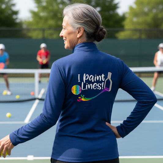 A woman standing on a pickleball court seen from behind, wearing a royal blue long-sleeve shirt with the phrase 'I paint Lines!' and a pickelball paddle graphic printed on the back including a multi-colored pickleball. 