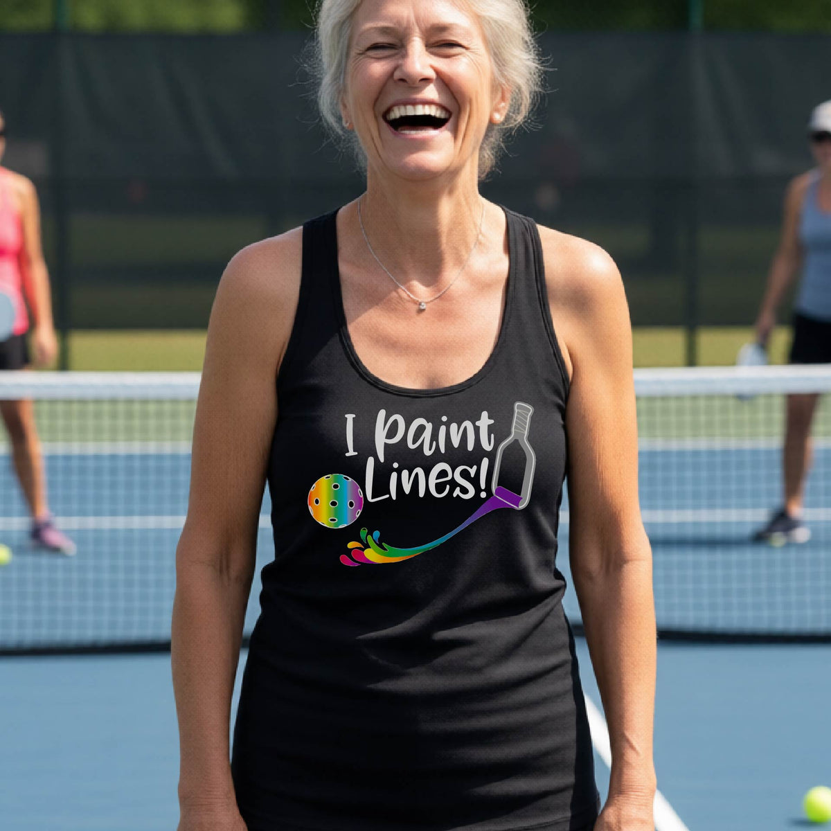 A close-up of a woman wearing a black racerback tank top with a graphic on the front that has white text "I PAINT LINES'  with a white pickleball paddle painting multi-colored lines leading a mutli-colored pickleball.