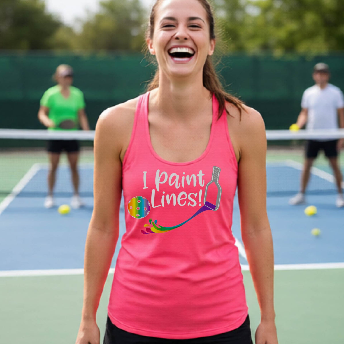 A close-up of a woman wearing a coral pink racerback tank top with a graphic on the front that has white text "I PAINT LINES'  with a white pickleball paddle painting multi-colored lines leading a mutli-colored pickleball.