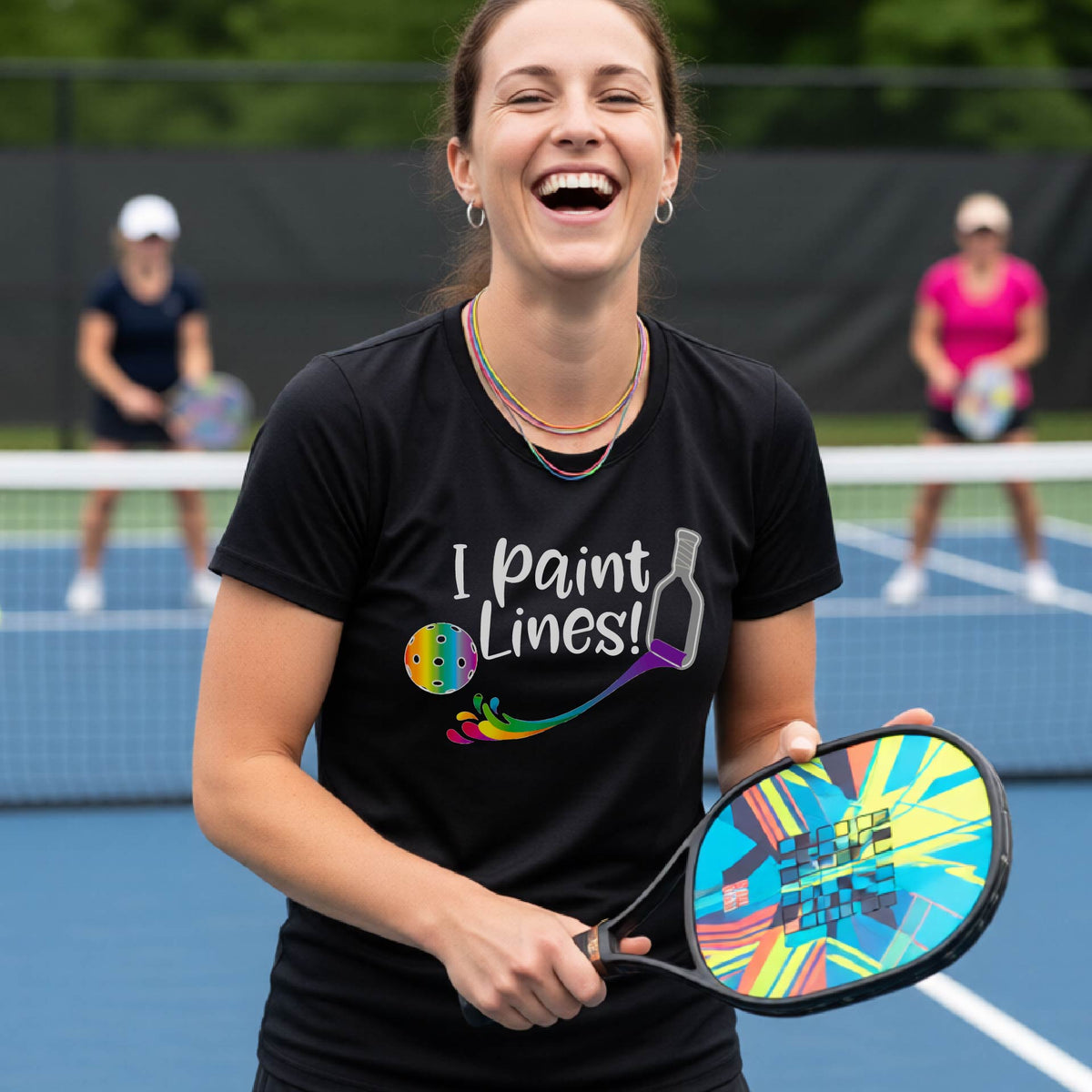 A close-up of a woman wearing a black short-sleeve t-shirt with a graphic on the front that has white text "I PAINT LINES'  with a white pickleball paddle painting multi-colored lines leading a mutli-colored pickleball.