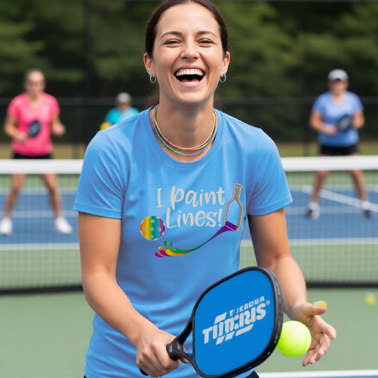 A close-up of a woman wearing a light blue short-sleeve t-shirt with a graphic on the front that has white text "I PAINT LINES'  with a white pickleball paddle painting multi-colored lines leading a mutli-colored pickleball.
