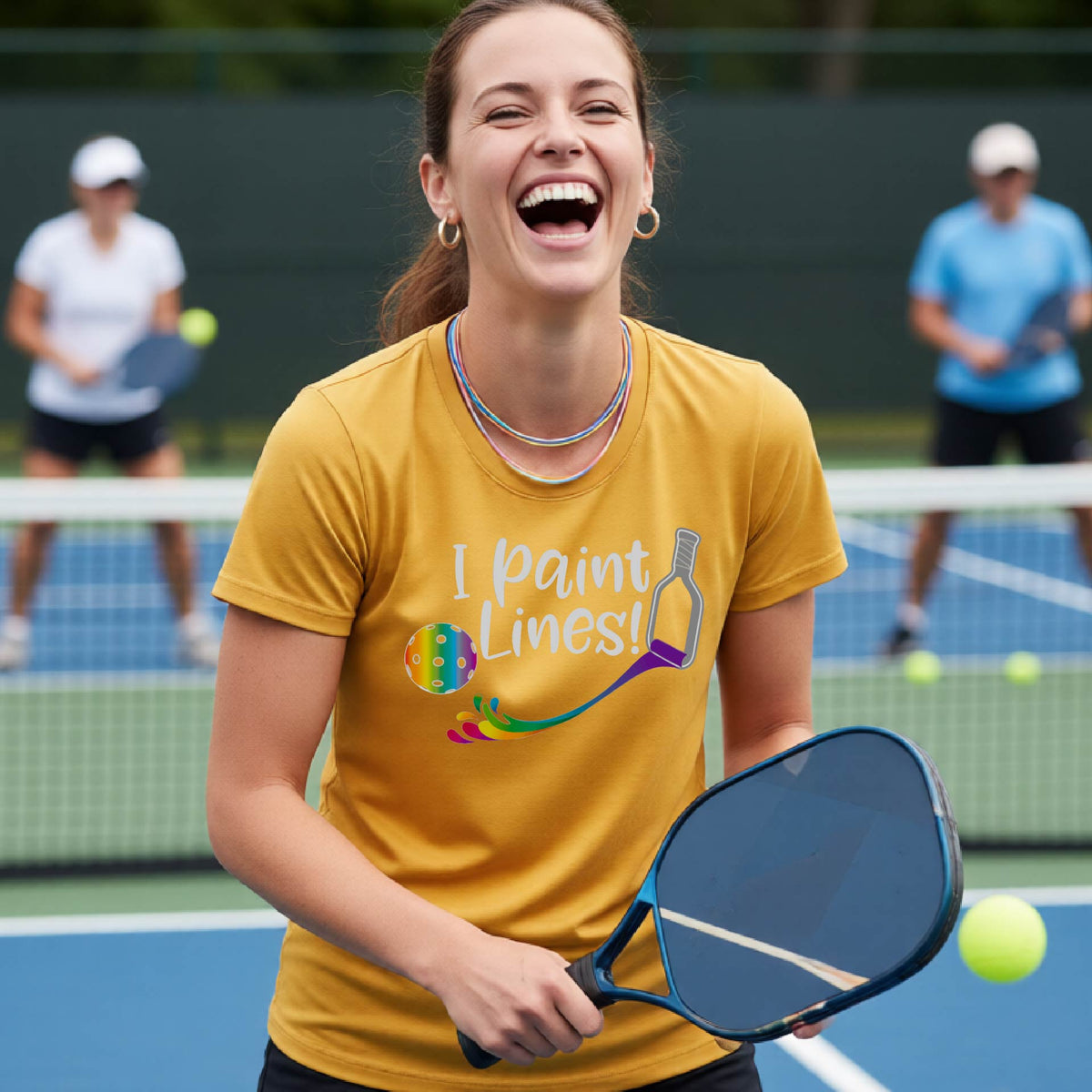 A close-up of a woman wearing a mustard yellow gold short-sleeve t-shirt with a graphic on the front that has white text "I PAINT LINES'  with a white pickleball paddle painting multi-colored lines leading a mutli-colored pickleball.