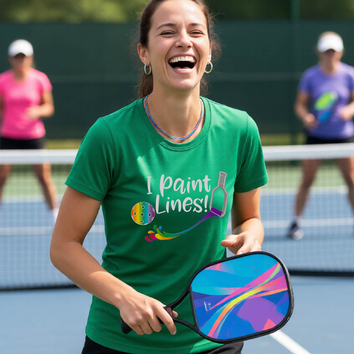 A close-up of a woman wearing a green short-sleeve t-shirt with a graphic on the front that has white text "I PAINT LINES'  with a white pickleball paddle painting multi-colored lines leading a mutli-colored pickleball.