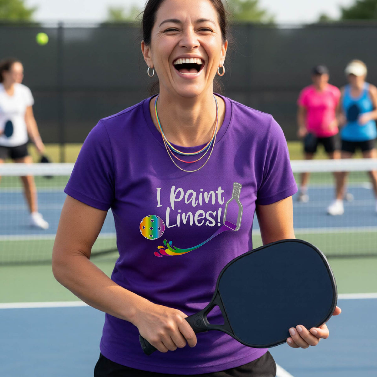 A close-up of a woman wearing a purple short-sleeve t-shirt with a graphic on the front that has white text "I PAINT LINES'  with a white pickleball paddle painting multi-colored lines leading a mutli-colored pickleball.