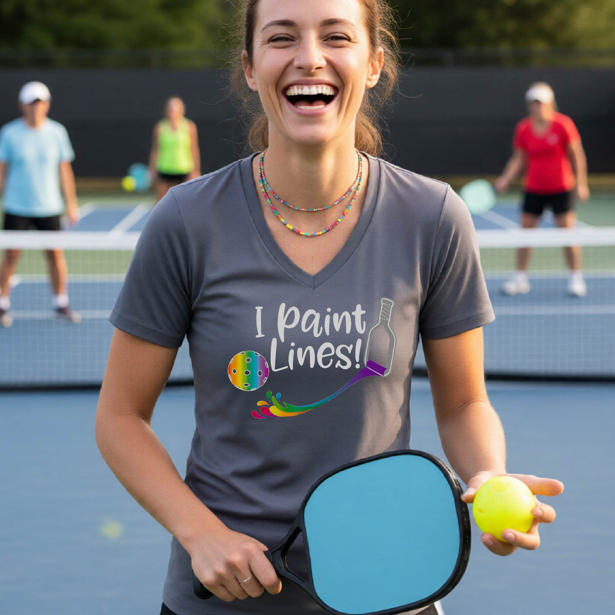 A woman on a pickleball court wearing a gray V-neck t-shirt with the graphic 'I paint Lines!' and a paintbrush illustration. 