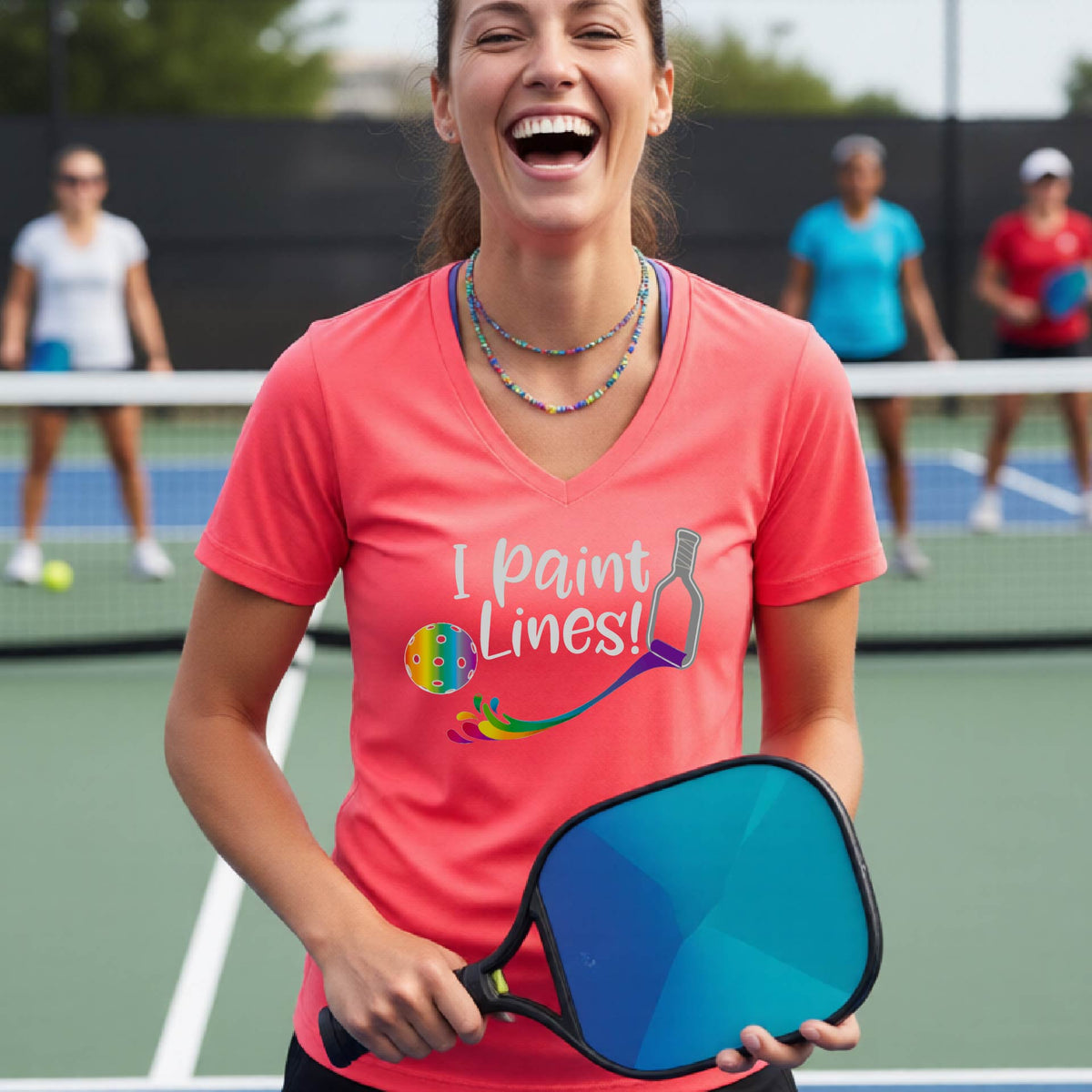 A woman on a pickleball court wearing a bright coral pink V-neck t-shirt with the graphic 'I paint Lines!' and a paintbrush illustration. 