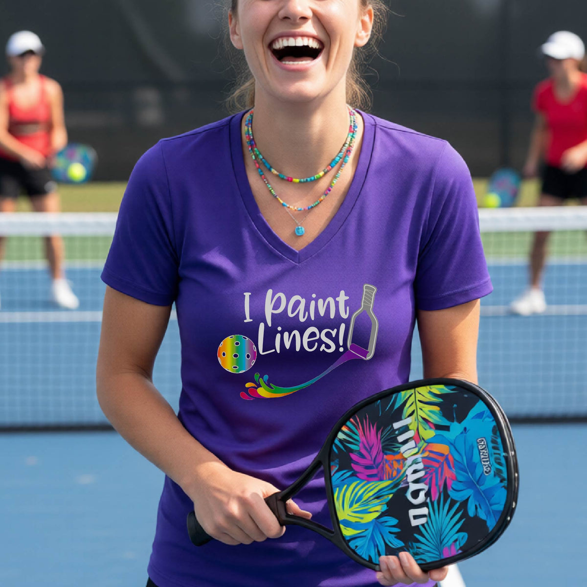 A woman on a pickleball court wearing a bright purple V-neck t-shirt with the graphic 'I paint Lines!' and a paintbrush illustration. 