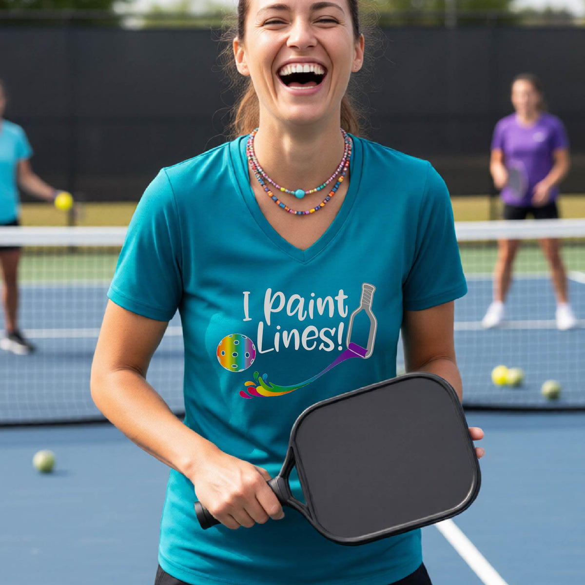 A woman on a pickleball court wearing a bright teal V-neck t-shirt with the graphic 'I paint Lines!' and a paintbrush illustration. 
