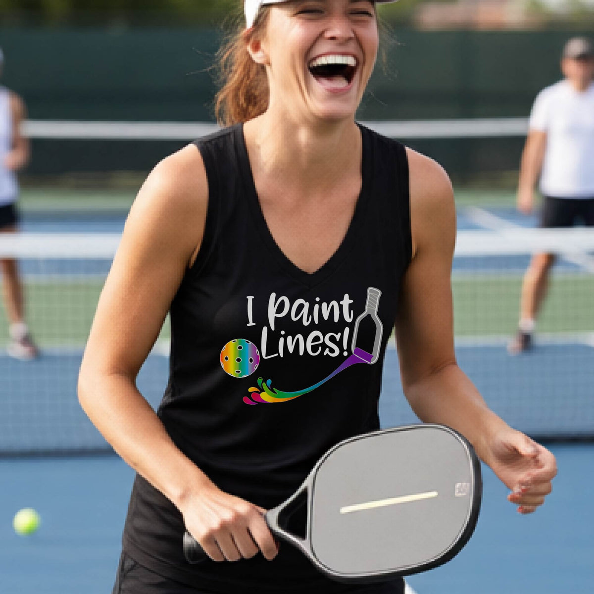 A woman laughing on a pickleball court wearing a black tank top with the text 'I paint lines!' and a paintbrush graphic, holding a paddle.