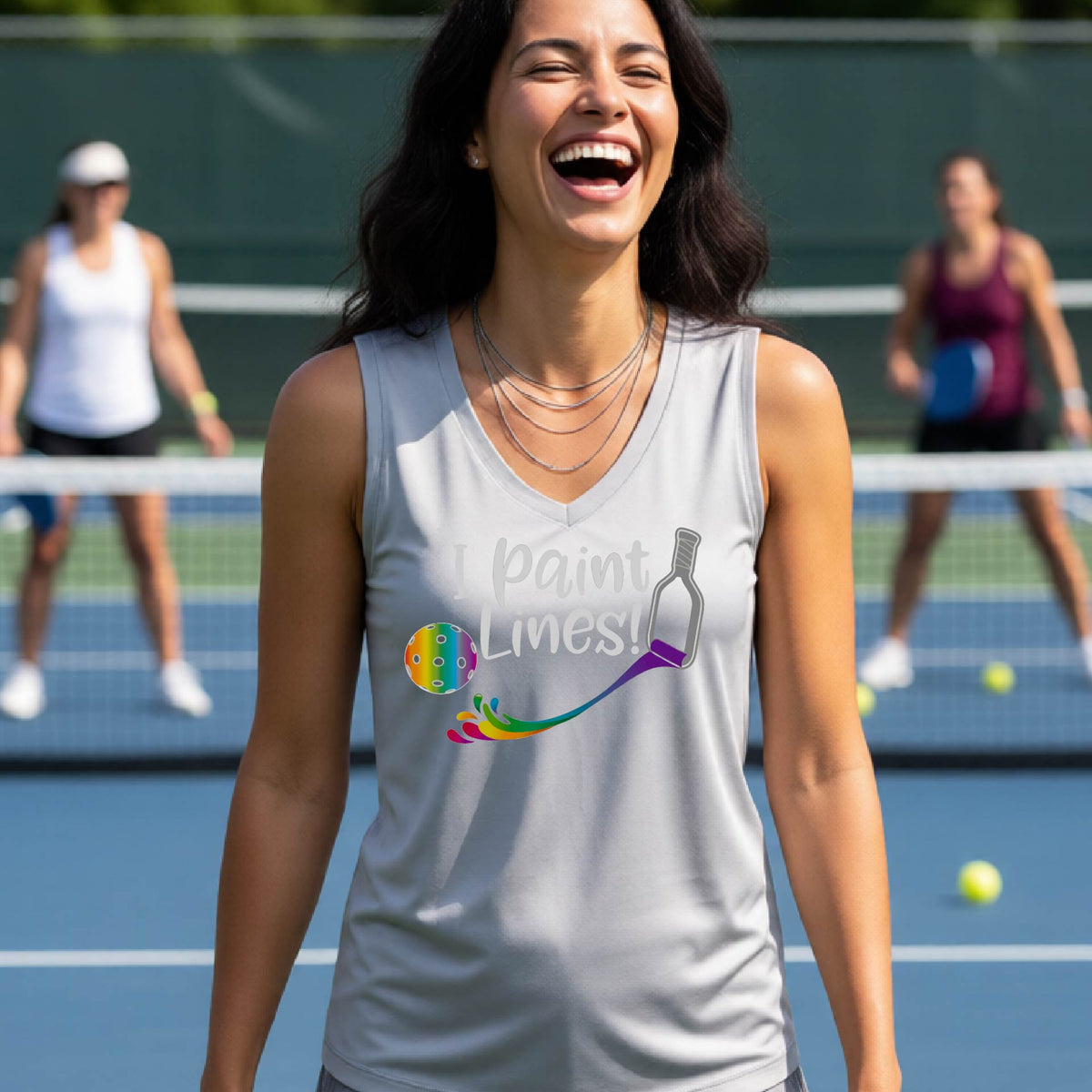 A close-up of a woman wearing a silver gray sleeveless t-shirt with a graphic on the front that has white text "I PAINT LINES'  with a white pickleball paddle painting multi-colored lines leading a mutli-colored pickleball.