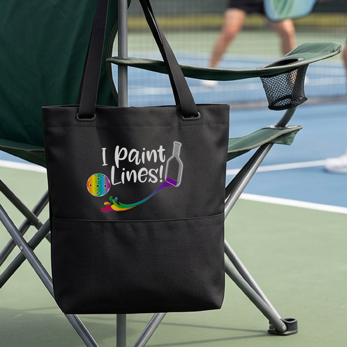 A close-up of black tote bag with a graphic on the front that has white text "I PAINT LINES'  with a white pickleball paddle painting multi-colored lines leading a mutli-colored pickleball.