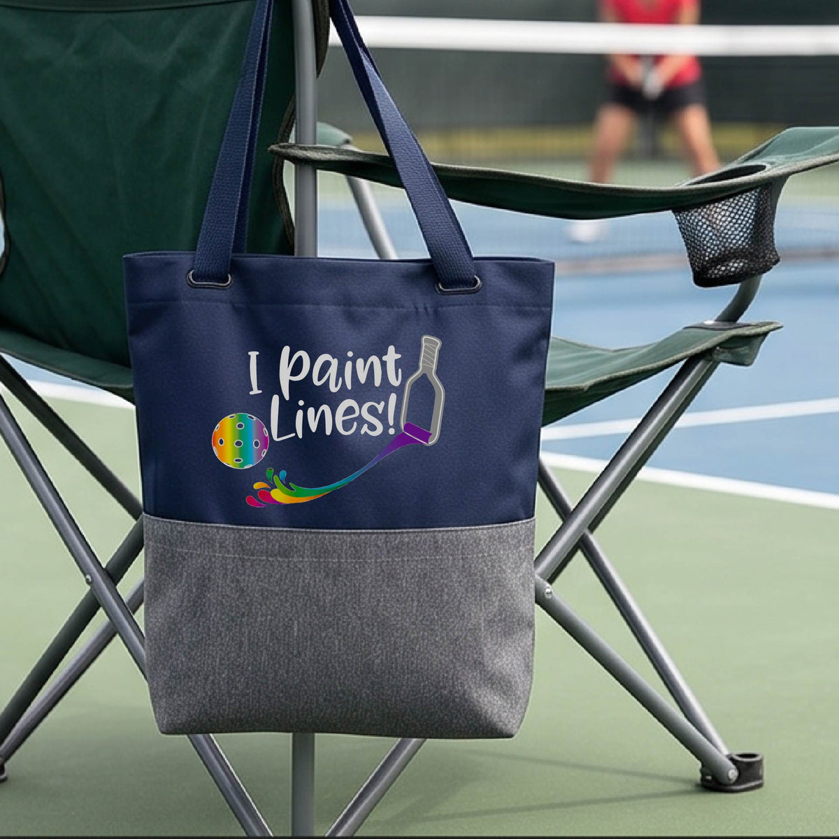 A close-up of blue and gray tote bag with a graphic on the front that has white text "I PAINT LINES'  with a white pickleball paddle painting multi-colored lines leading a mutli-colored pickleball.