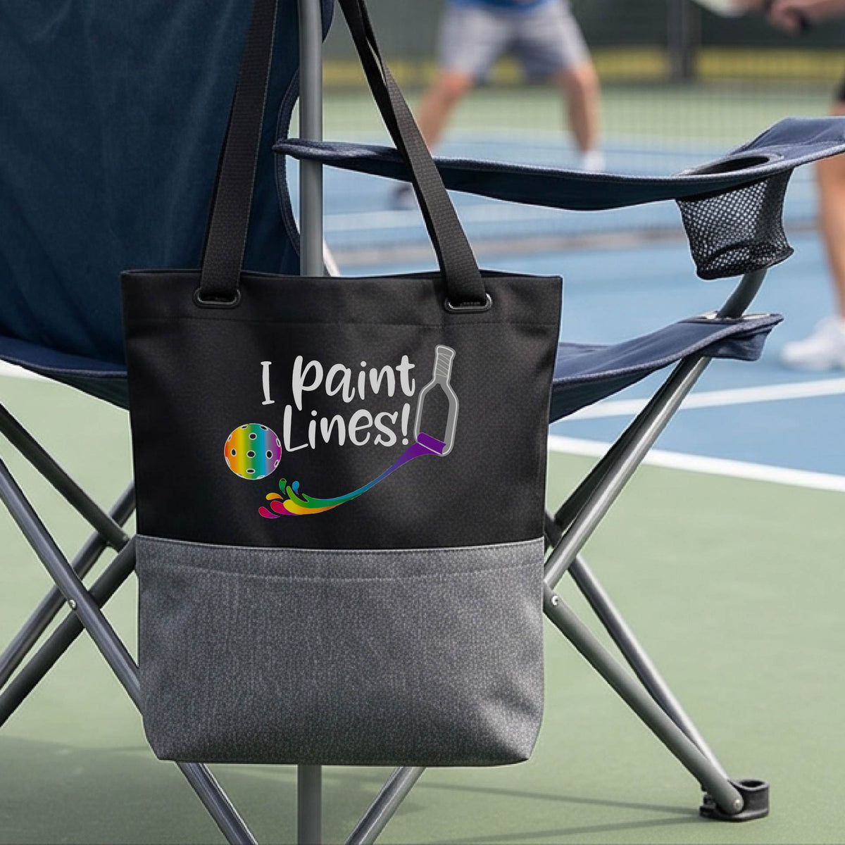 A close-up of black and gray tote bag with a graphic on the front that has white text "I PAINT LINES'  with a white pickleball paddle painting multi-colored lines leading a mutli-colored pickleball.