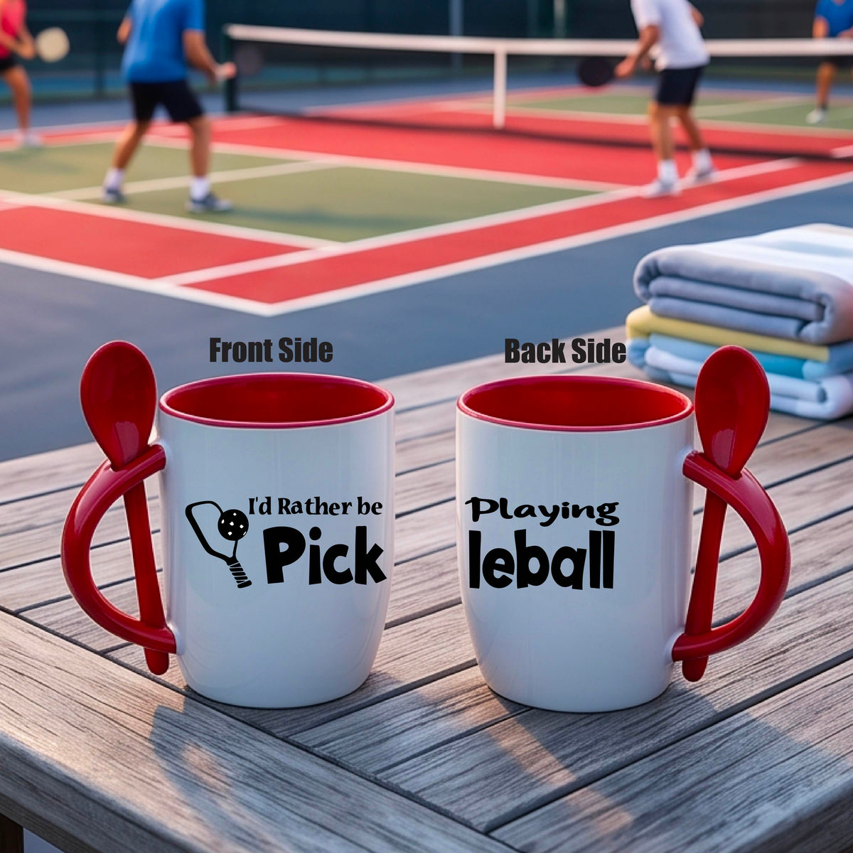 A white and red 12 ounce coffee mug with a built in spoon in the handle with and a fun pickleball paddle and ball graphic and the text “I’D RATHER BE PLAYING PICKELBALL”