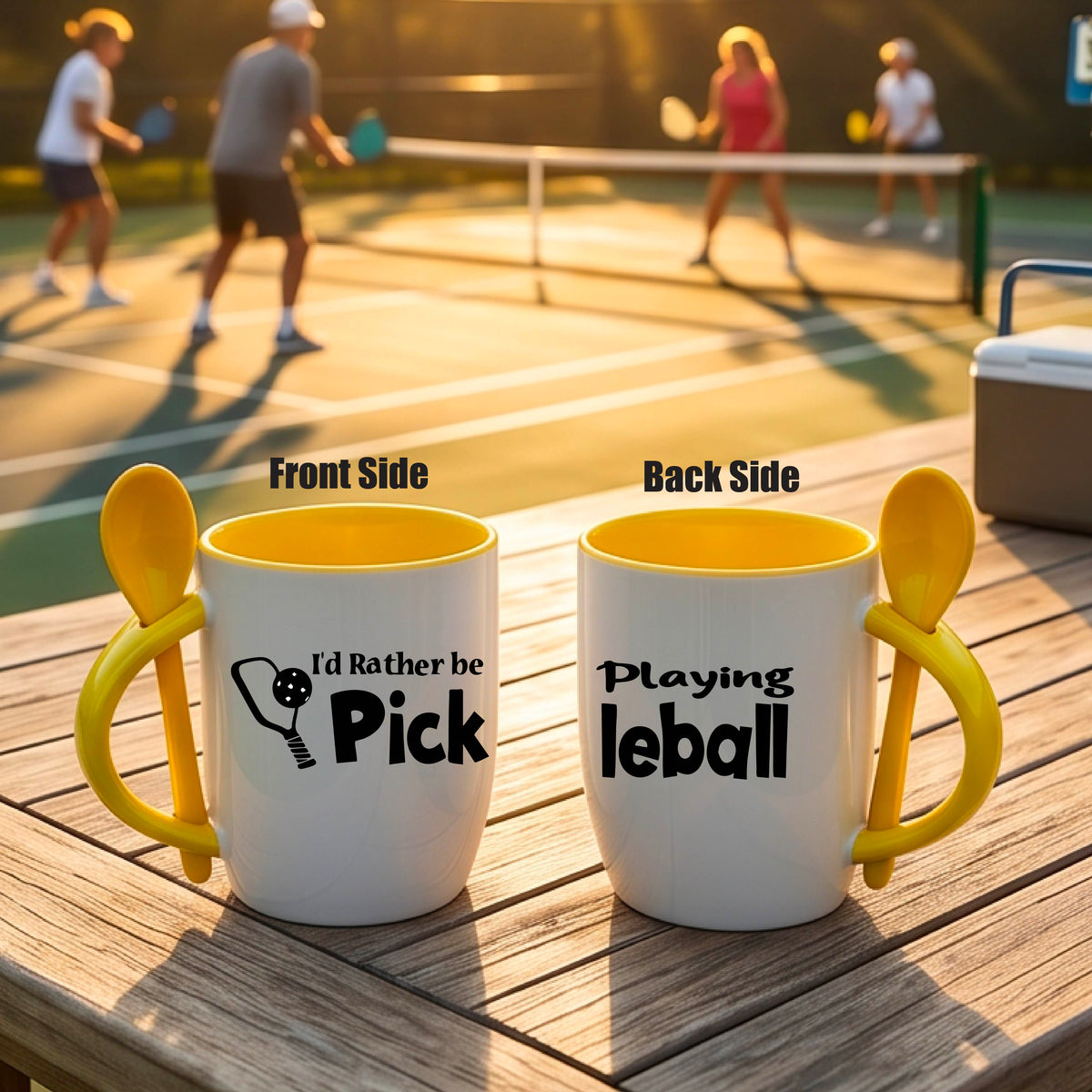 A white and yellow 12 ounce coffee mug with a built in spoon in the handle with and a fun pickleball paddle and ball graphic and the text “I’D RATHER BE PLAYING PICKELBALL”