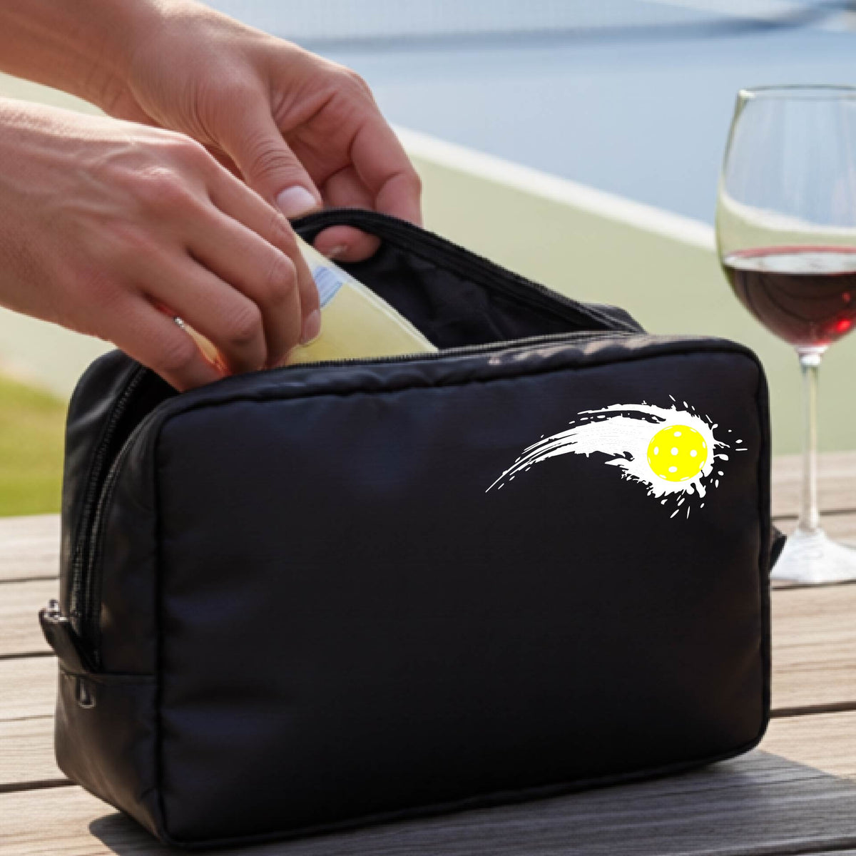 A close-up of a black cosmetic toiletry bag with a white pickleball graphic of a yellow pickleball with a motion trail effect, displayed in front of a pickleball court. 