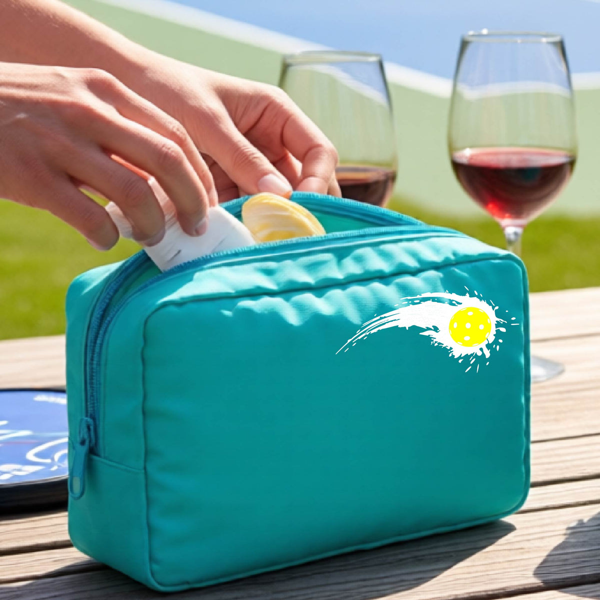A close-up of a turquoise cosmetic toiletry bag with a white pickleball graphic of a yellow pickleball with a motion trail effect, displayed in front of a pickleball court. 