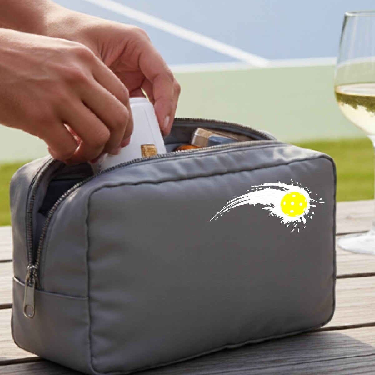 A close-up of a gray cosmetic toiletry bag with a white pickleball graphic of a yellow pickleball with a motion trail effect, displayed in front of a pickleball court. 