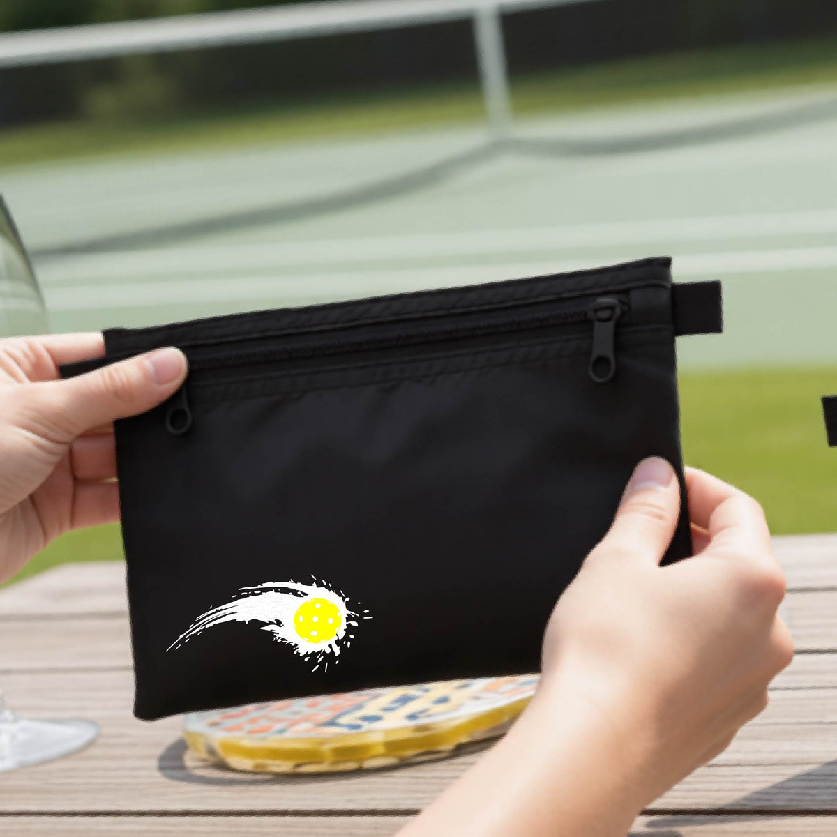 A close-up of a black cosmetic toiletry bag with a white pickleball graphic of a yellow pickleball with a motion trail effect, displayed in front of a pickleball court. 

