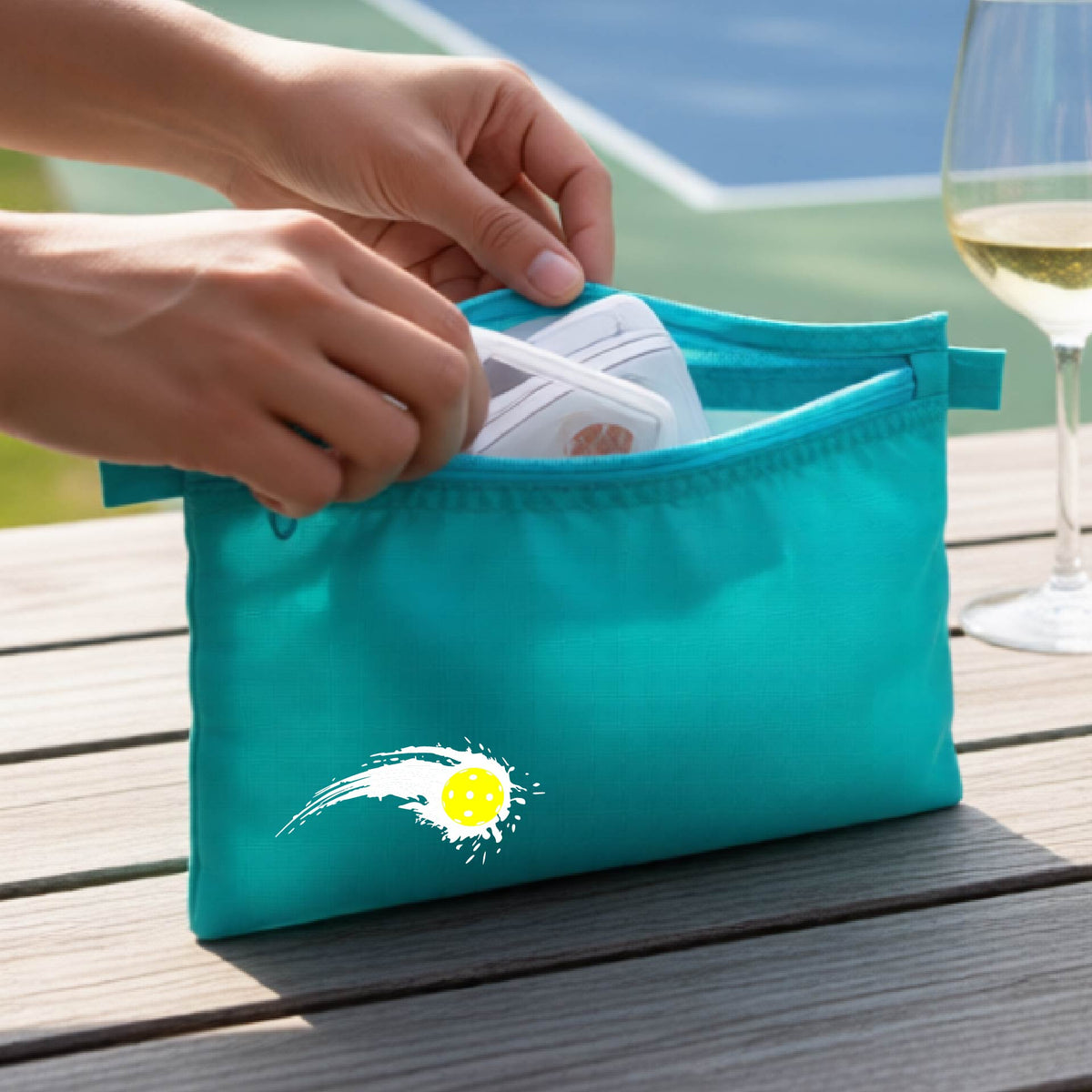 A close-up of a turquoise cosmetic toiletry bag with a white pickleball graphic of a yellow pickleball with a motion trail effect, displayed in front of a pickleball court.