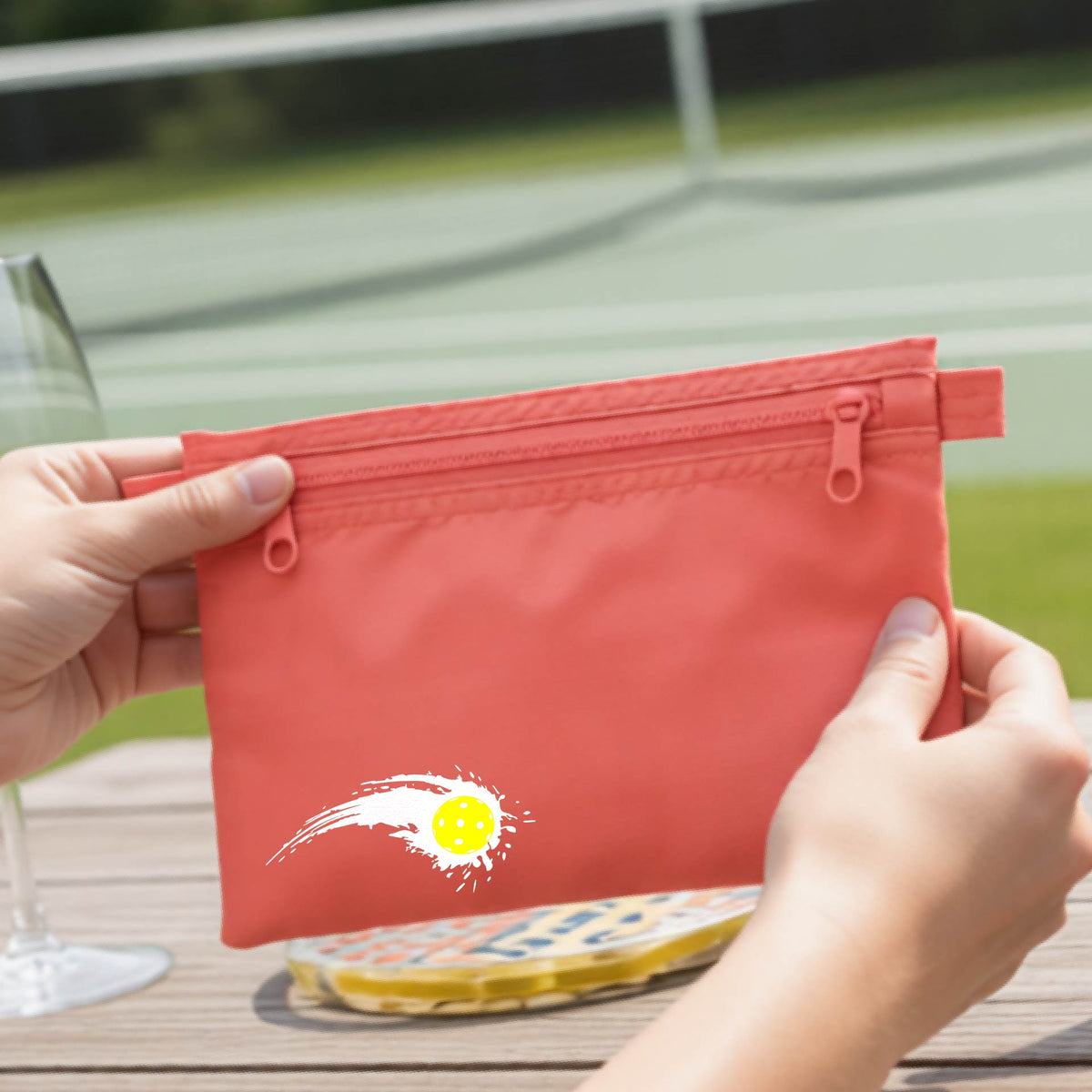 A close-up of a coral colored cosmetic toiletry bag with a white pickleball graphic of a yellow pickleball with a motion trail effect, displayed in front of a pickleball court.