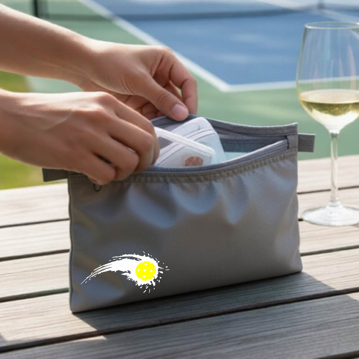 A close-up of a gray cosmetic toiletry bag with a white pickleball graphic of a yellow pickleball with a motion trail effect, displayed in front of a pickleball court.