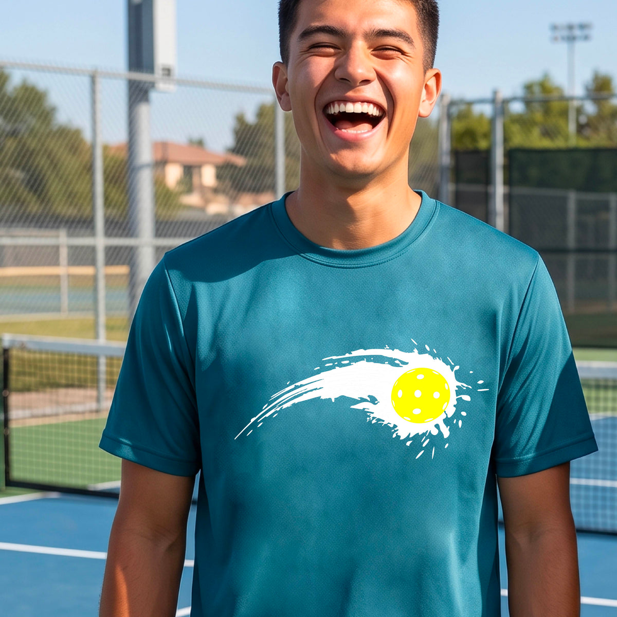 A man wearing a teal short-sleeve T-shirt with a white pickleball graphic of a yellow pickleball with a motion trail effect, displayed in front of a pickleball court. 