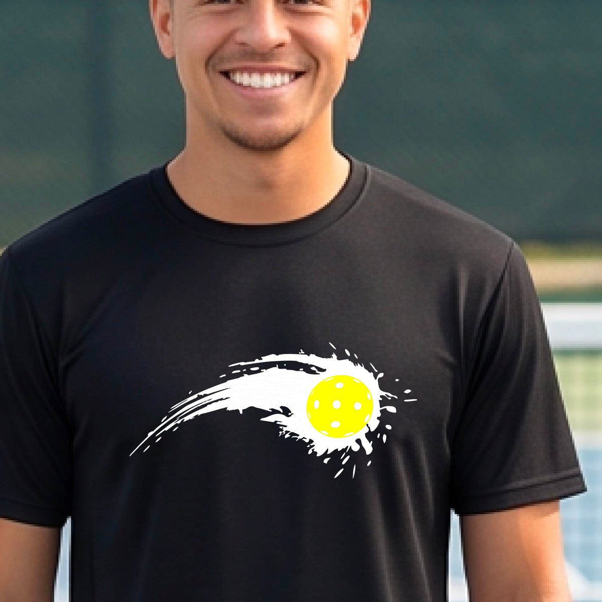 A man wearing a black short-sleeve T-shirt with a white pickleball graphic of a yellow pickleball with a motion trail effect, displayed in front of a pickleball court. 