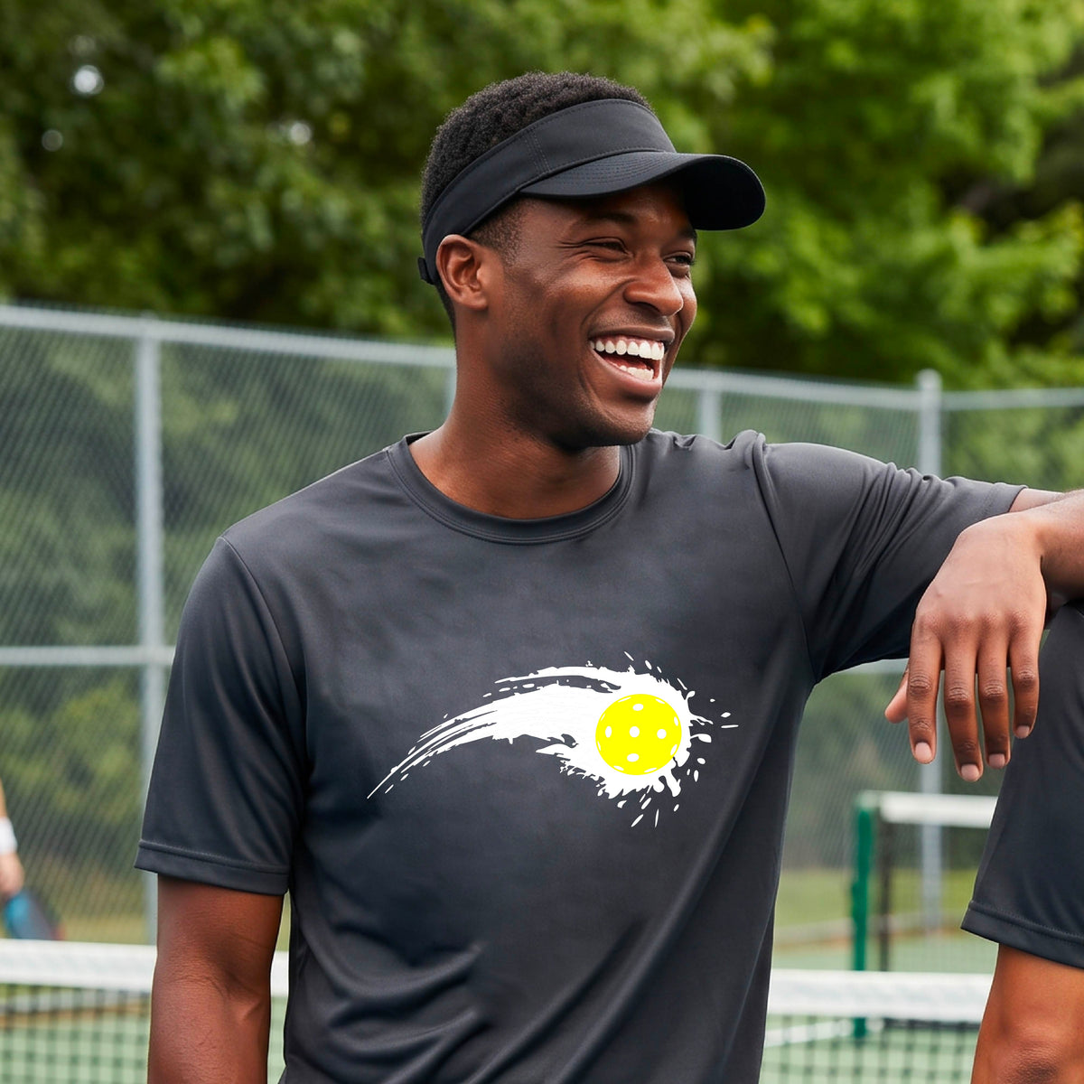A man wearing a dark gray short-sleeve T-shirt with a white pickleball graphic of a yellow pickleball with a motion trail effect, displayed in front of a pickleball court. 