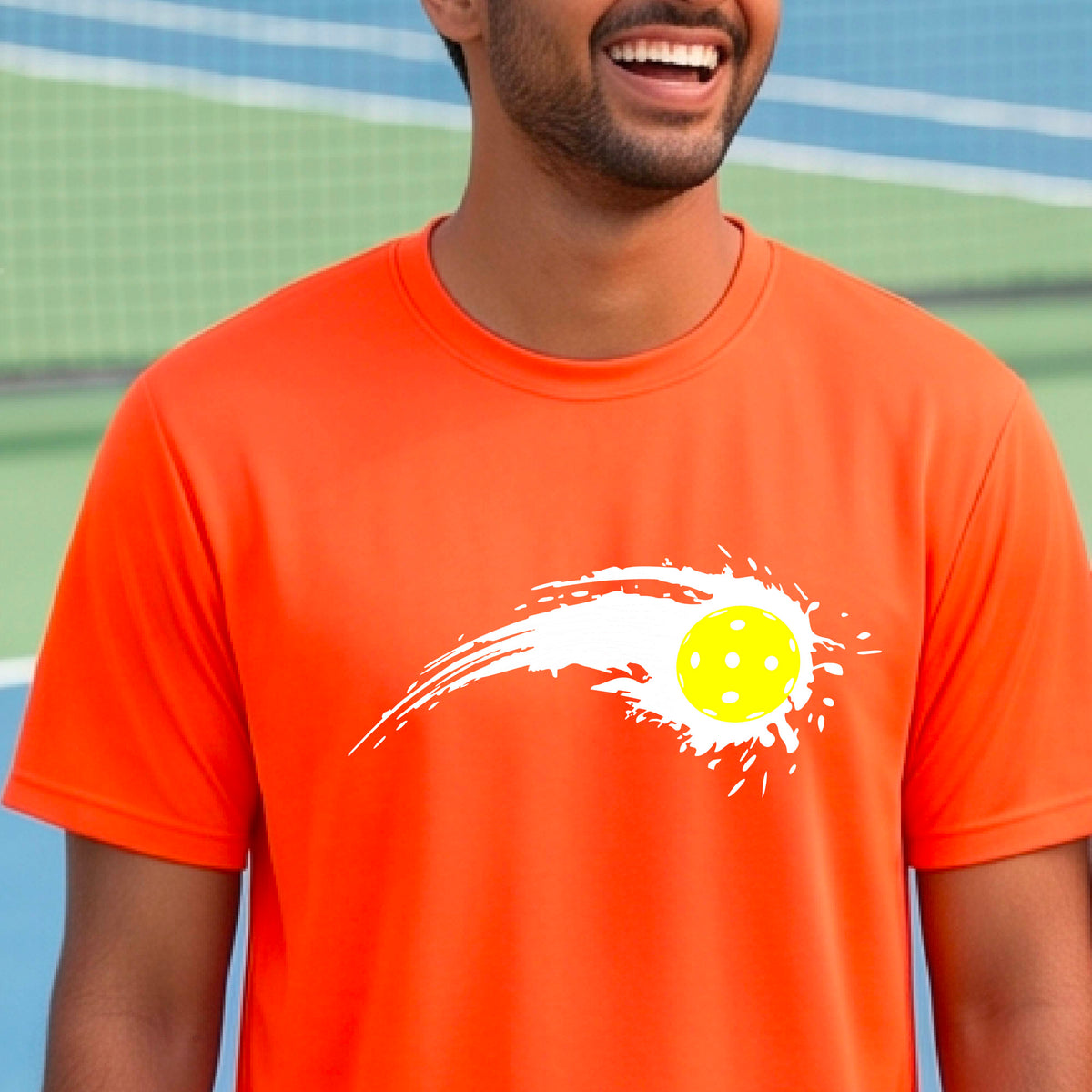 A man wearing an orange short-sleeve T-shirt with a white pickleball graphic of a yellow pickleball with a motion trail effect, displayed in front of a pickleball court. 