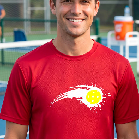 A man wearing a red short-sleeve T-shirt with a white pickleball graphic of a yellow pickleball with a motion trail effect, displayed in front of a pickleball court. 