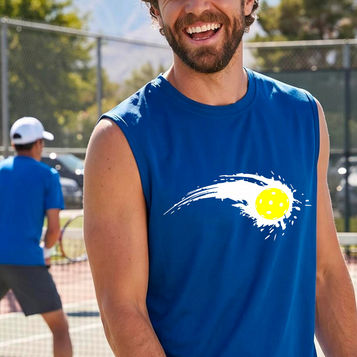 A man wearing a royal blue sleeveless T-shirt with a white pickleball graphic of a yellow pickleball with a motion trail effect, displayed in front of a pickleball court. 