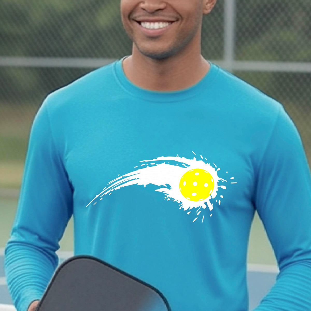 A man wearing a bright blue long-sleeve T-shirt with a white pickleball graphic of a yellow pickleball with a motion trail effect, displayed in front of a pickleball court. 