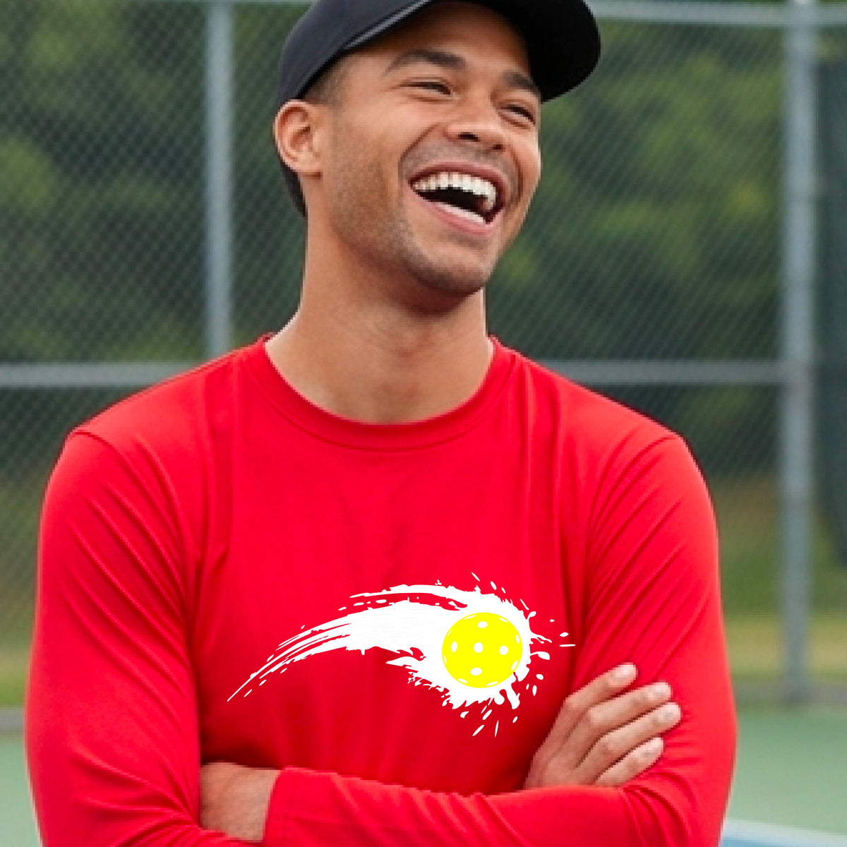 A man wearing a red long-sleeve T-shirt with a white pickleball graphic of a yellow pickleball with a motion trail effect, displayed in front of a pickleball court. 