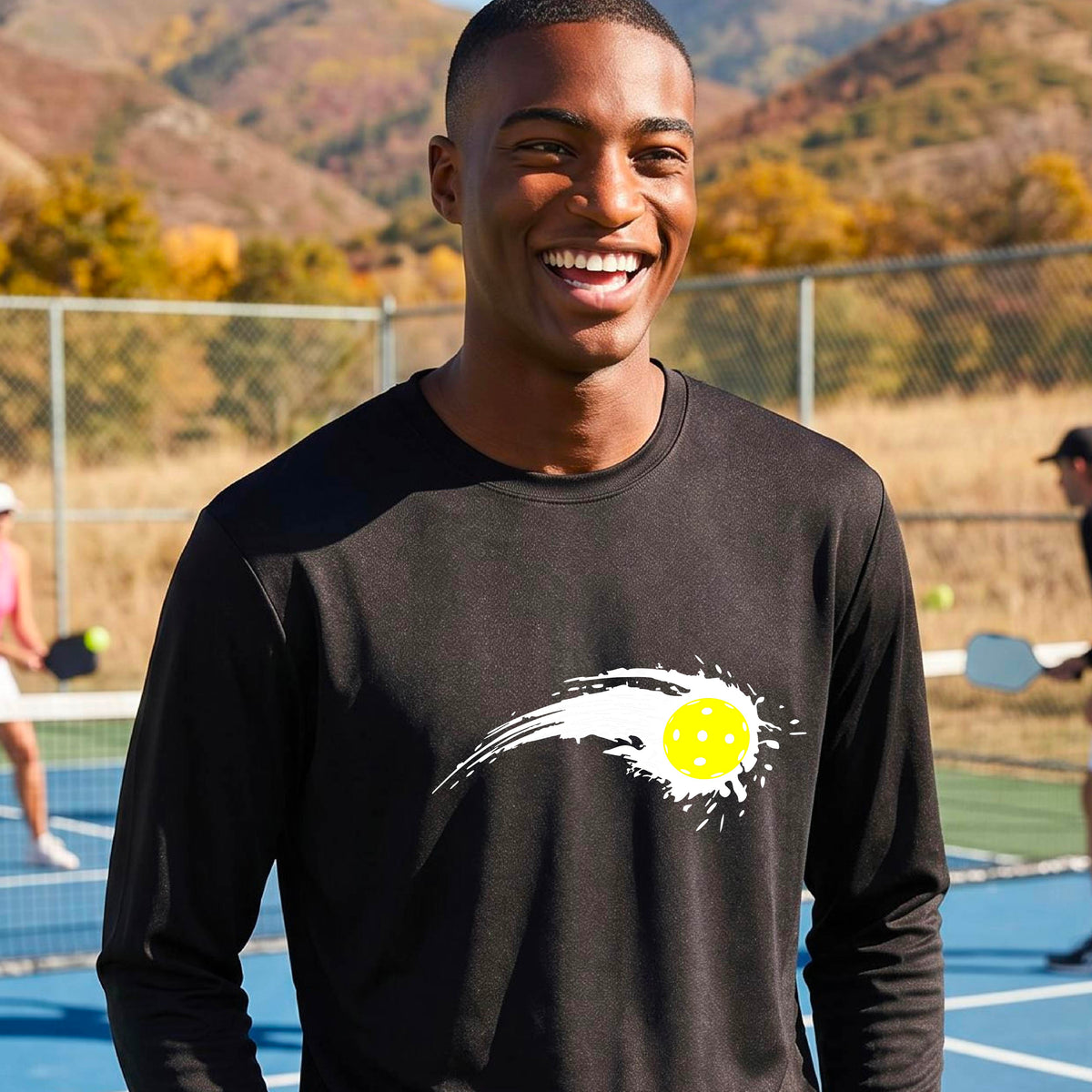 A man wearing a black long-sleeve T-shirt with a white pickleball graphic of a yellow pickleball with a motion trail effect, displayed in front of a pickleball court. 