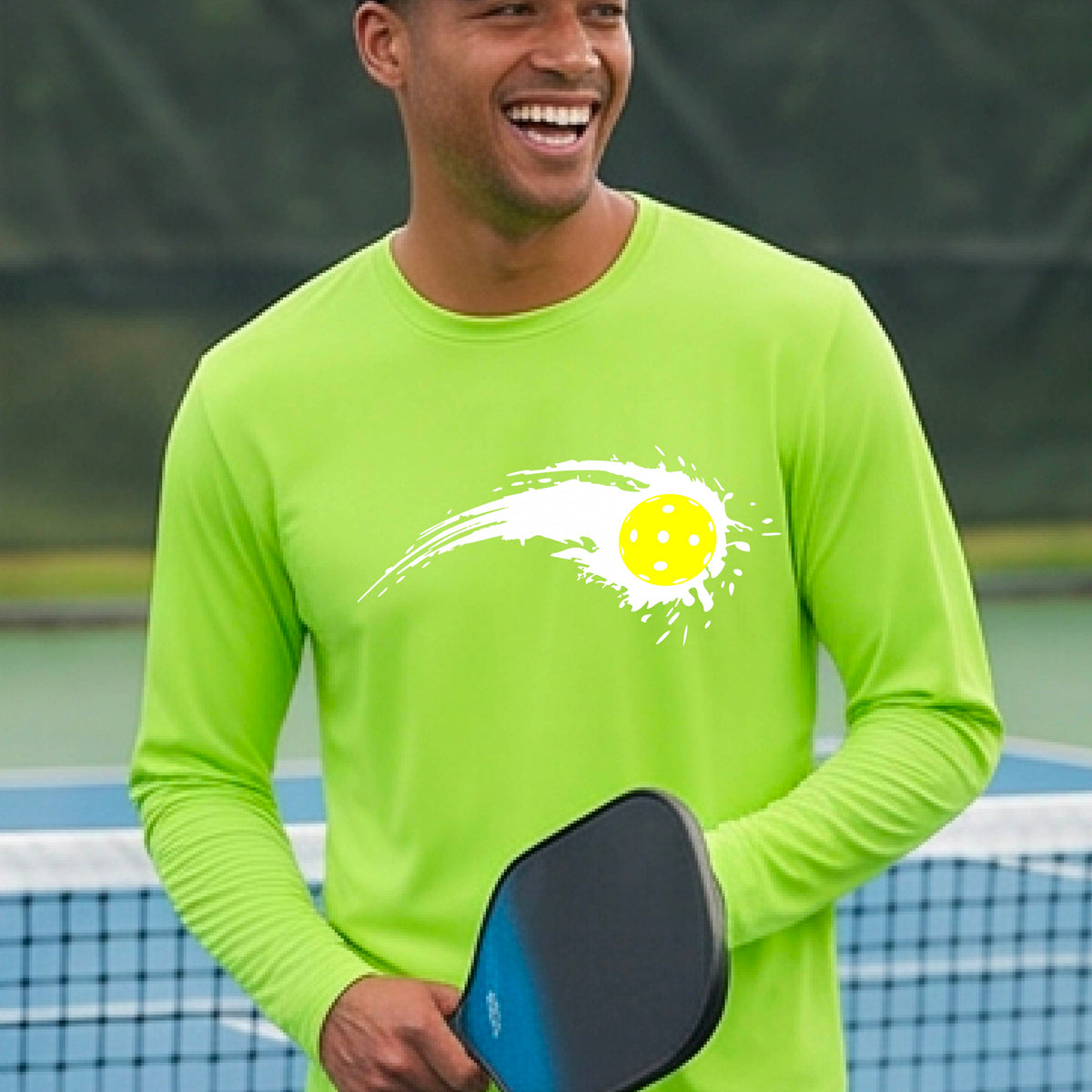 A man wearing a bright lime green long-sleeve T-shirt with a white pickleball graphic of a yellow pickleball with a motion trail effect, displayed in front of a pickleball court. 