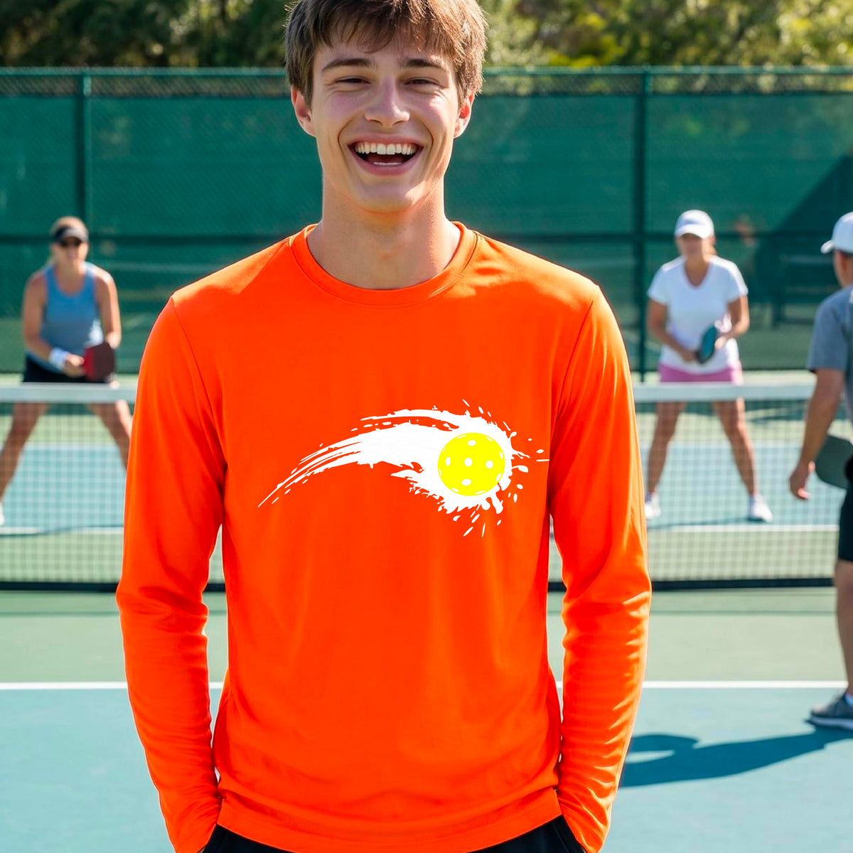 A man wearing a bright orange long-sleeve T-shirt with a white pickleball graphic of a yellow pickleball with a motion trail effect, displayed in front of a pickleball court. 