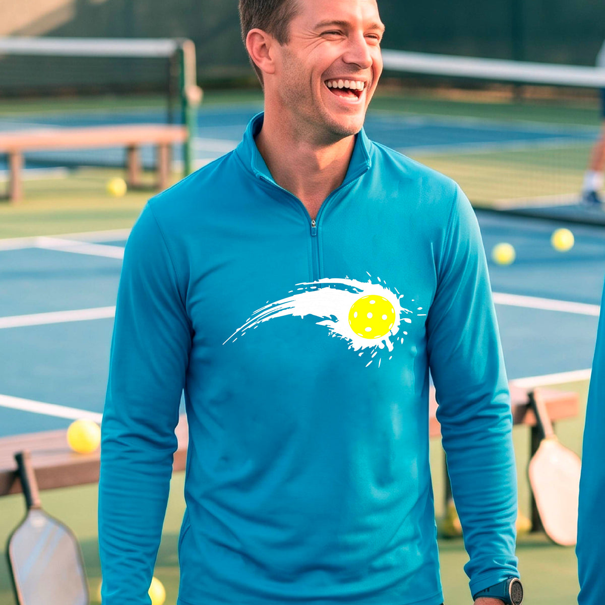 A man wearing a blue long-sleeve quarter-zip shirt with a white pickleball graphic of a yellow pickleball with a motion trail effect, displayed in front of a pickleball court. 