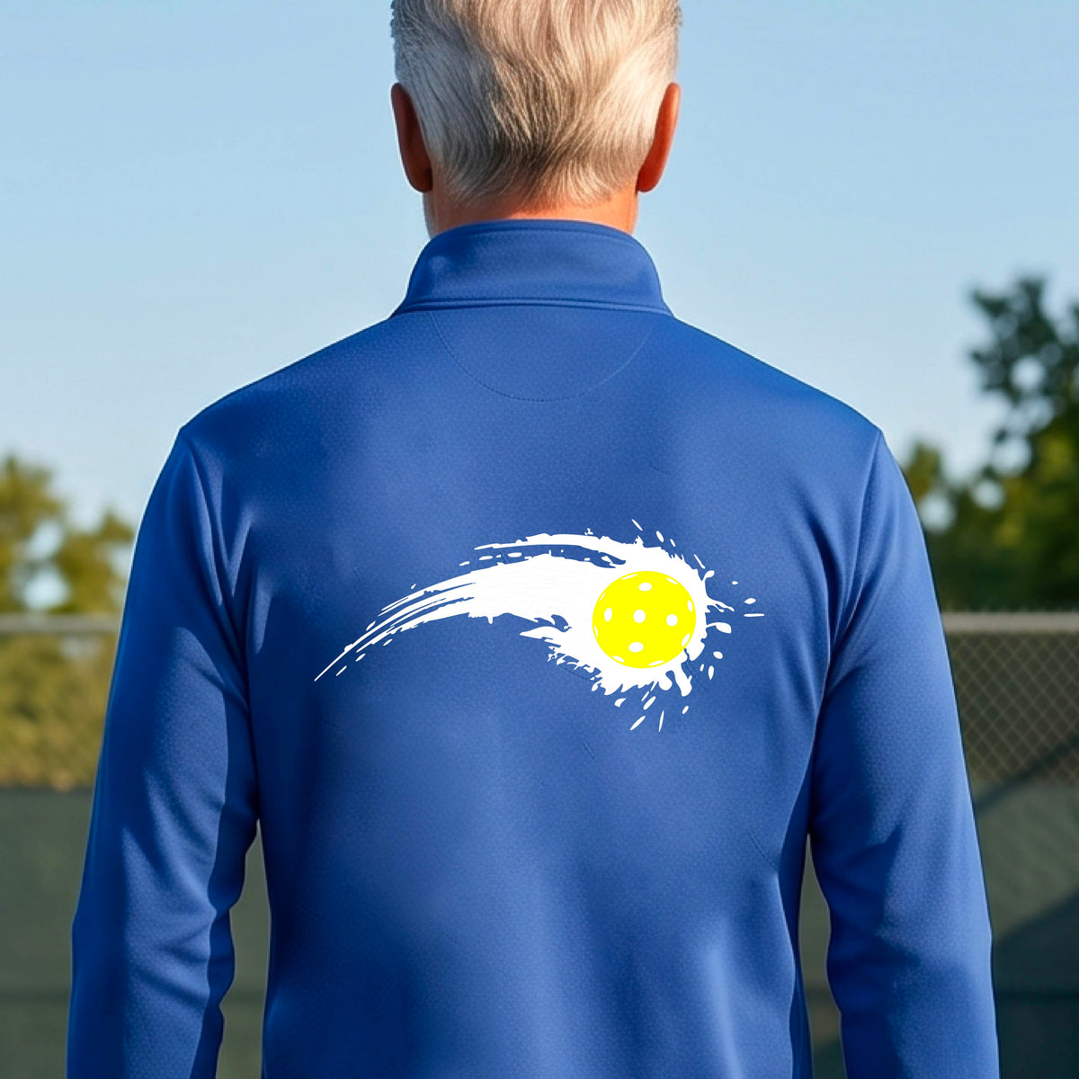 A man wearing a royal blue long-sleeve quarter-zip shirt with a white pickleball graphic of a yellow pickleball with a motion trail effect, displayed in front of a pickleball court. 