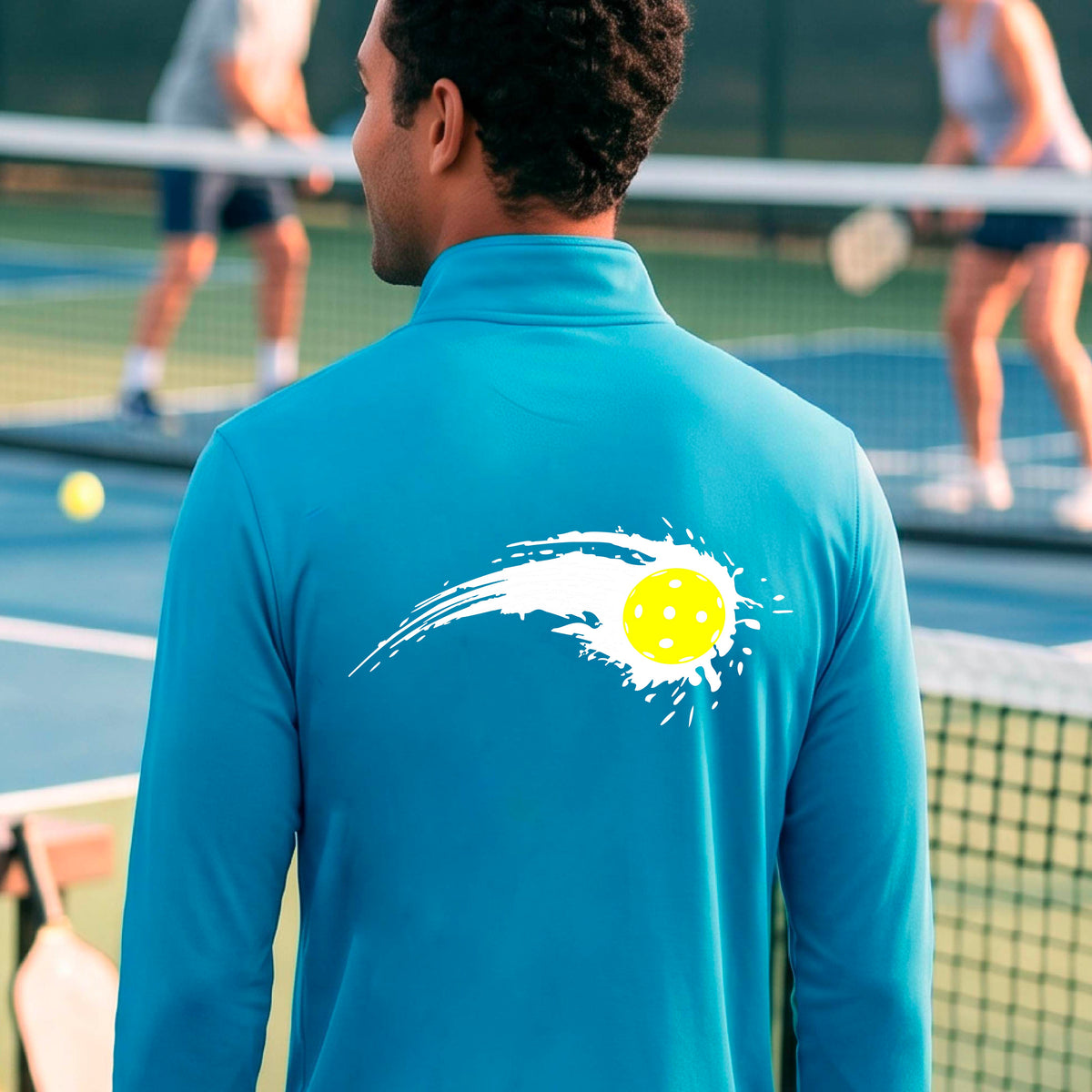 A man wearing a blue long-sleeve quarter-zip shirt with a white pickleball graphic of a yellow pickleball with a motion trail effect, displayed in front of a pickleball court. 