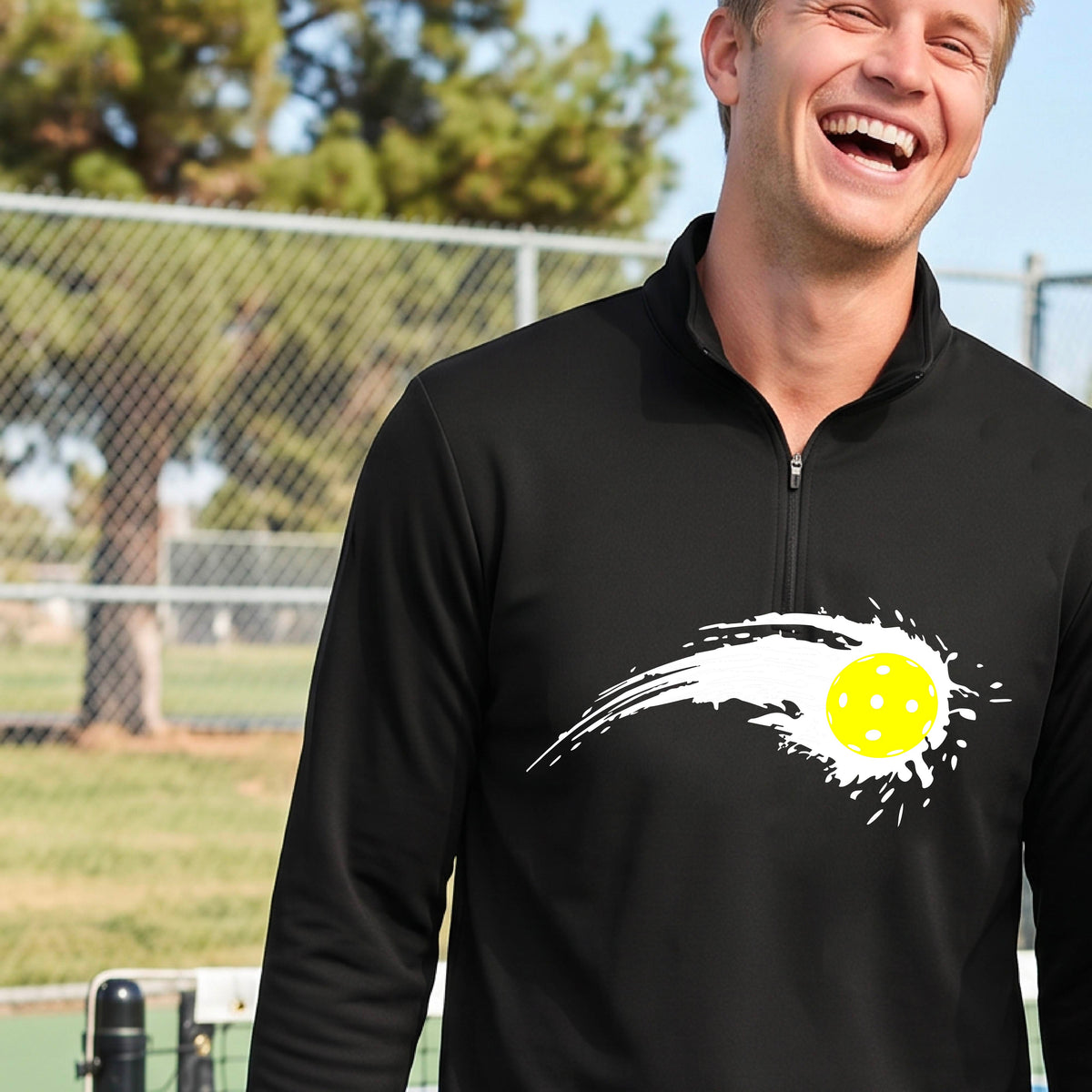 A man wearing a black long-sleeve quarter-zip shirt with a white pickleball graphic of a yellow pickleball with a motion trail effect, displayed in front of a pickleball court. 