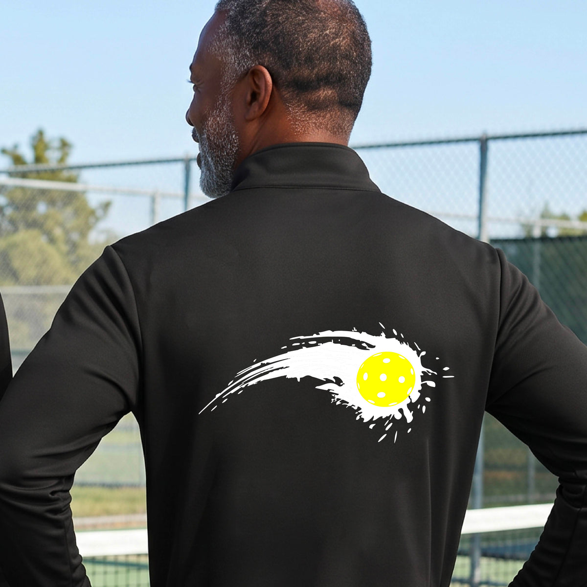 A man wearing a black long-sleeve quarter-zip shirt with a white pickleball graphic of a yellow pickleball with a motion trail effect, displayed in front of a pickleball court. 