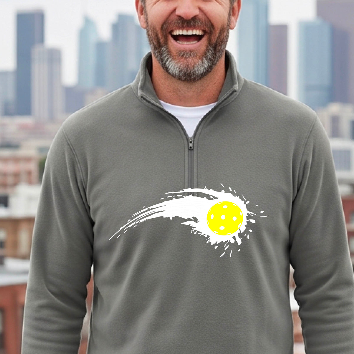 A man wearing a gray long-sleeve quarter-zip shirt with a white pickleball graphic of a yellow pickleball with a motion trail effect, displayed in front of a pickleball court. 