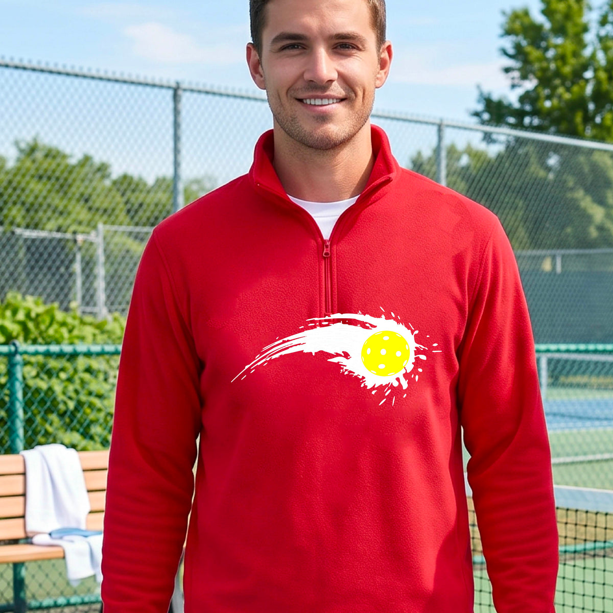 A man wearing a red long-sleeve quarter-zip shirt with a white pickleball graphic of a yellow pickleball with a motion trail effect, displayed in front of a pickleball court. 